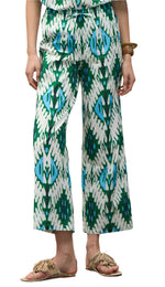 A person stands against a white background wearing the Figue Justine Pant—white with green and blue abstract patterns, side pockets, and a drawstring waist—paired with tan fringe sandals and a brown bracelet.