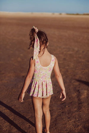 A young girl wearing Louise Misha's floral Baby Girls' and Girls' Kalinda Bathing Suit strolls alone on a sandy beach beneath a clear blue sky with her hair tied using a matching ribbon.