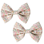 The Milledeux Girls' Large Bowtie Bow in Liberty Katie & Millie B fabric features charming floral patterns in pink, green, and orange on white, secured with grosgrain-covered alligator clips for easy styling.