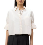 A person stands against a plain background wearing the TWP Next Ex Shirt, a vintage-washed poplin cropped button-down with cuffed sleeves and a large front pocket, paired with dark high-waisted pants.