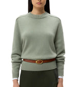 Someone wears the TWP New Jill Crewneck Sweater in light green Italian cashmere, featuring ribbed cuffs and hem, belted at the waist with a brown belt, and styled with dark pants.