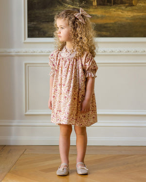 A young girl with curly hair wears the Noralee Girls' Maddie Dress, featuring vintage-inspired floral embroidery, paired with a matching bow and light shoes, as she stands indoors looking to the side.