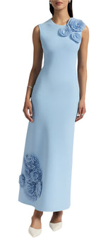 A woman stands against a plain background wearing Lela Rose's light blue, sleeveless knit midi dress with an A-line silhouette and floral shoulder and hem appliqués, paired with white pointed heels.
