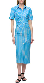 A woman wears the Lou Dress by Callas Milano, a blue and white vertical striped Italian cotton twill shirt dress with short sleeves and midi length, styled with black sandals against a white background.