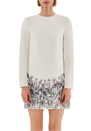 A person is wearing the Tilley Silk Wool Dress by Adam Lippes, an ivory ensemble featuring long sleeves and a charming floral print near the hem against a subtle background.