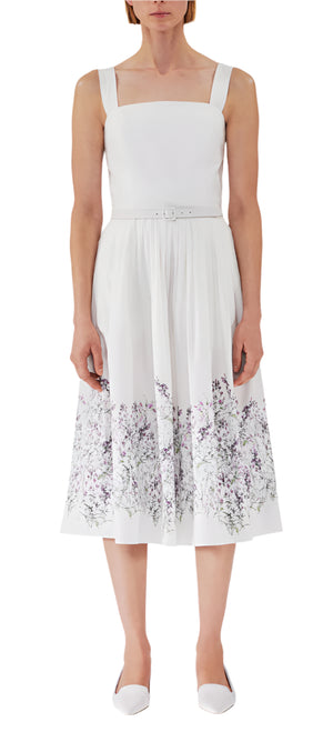 A person in an Adam Lippes Hoban Dress, featuring white poplin and purple floral prints, pairs it with white shoes against a plain backdrop.