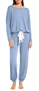 A woman stands barefoot wearing the Eberjey EBJ Gisele Slouchy Set—a loose-fitting, long-sleeve light blue pajama set with cuffed ankles and a white waist tie, made from sustainable TENCEL™ Modal.