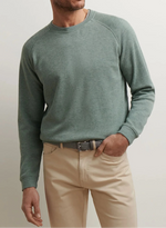 A person wearing the Peter Millar Crown Comfort Knit Crew in sage green, paired with beige pants and a black belt, stands with one hand in their pocket.
