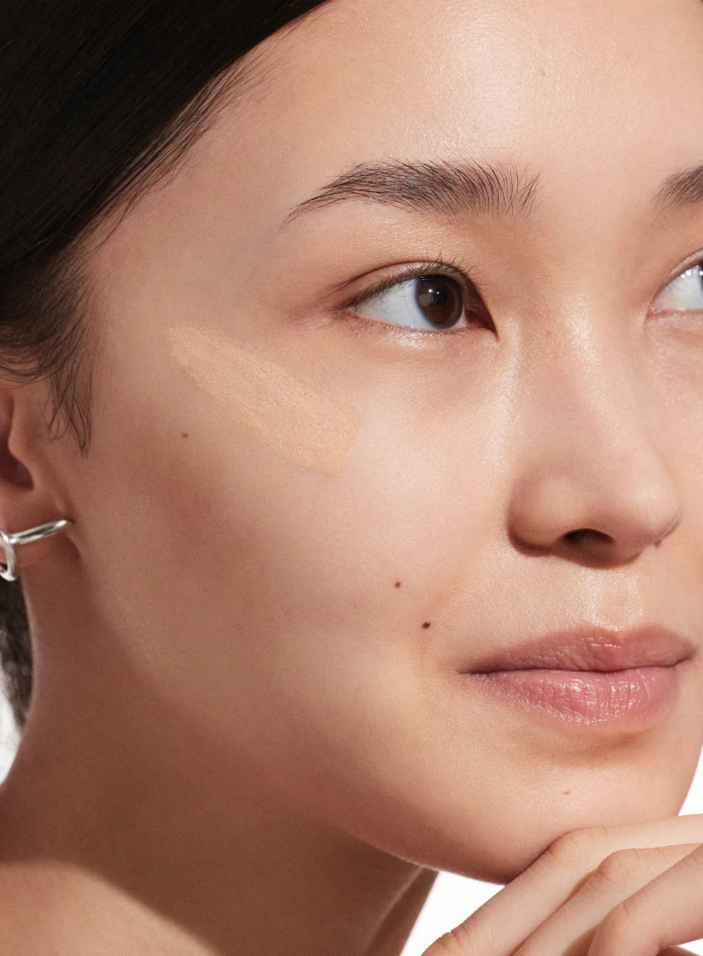 A woman with clear skin and a silver ear cuff rests her chin on her hand, gazing upward. A swatch of Supergoop Mineral Mattescreen SPF 40, a pore-blurring mattifying tinted sunscreen, is applied to her cheek.