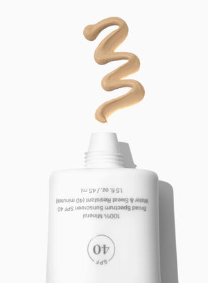 A white tube of Supergoop Mineral Mattescreen SPF 40 dispenses a swirl of beige mineral sunscreen on a white background, showcasing its mattifying, tinted formula that blurs pores.