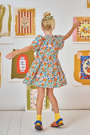 A young girl with blonde hair in a bun wears the Hunter Bell NYC Girls' Merritt Dress featuring a ruffled neckline, paired with yellow socks and mismatched sandals, standing with arms outstretched before framed wall art.