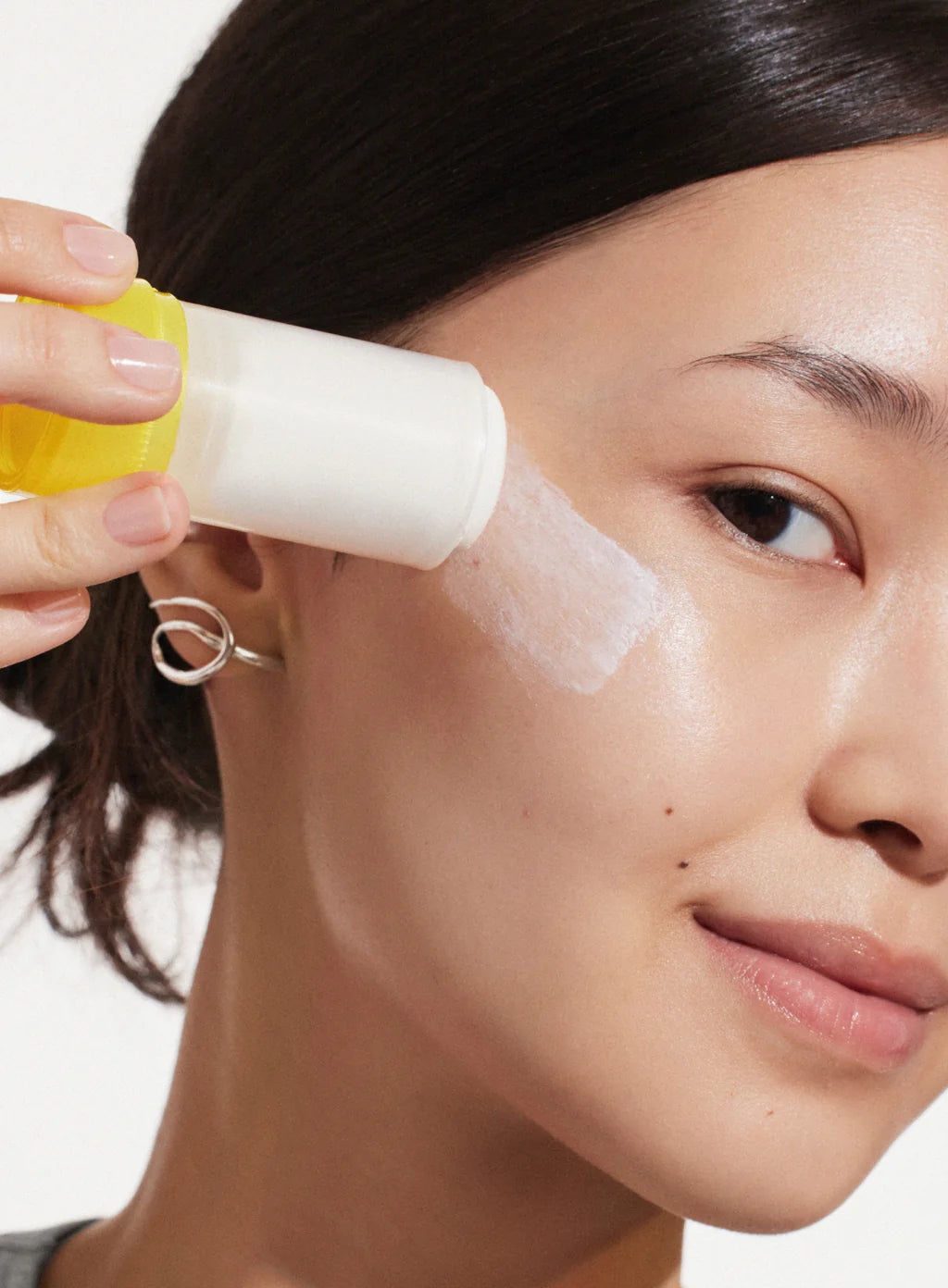 A person applies Supergoop Mineral Sheer Stick SPF 30 to their cheek, holding the yellow-capped applicator close to their face.