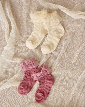 Two pairs of Noralee Ruffle Socks for children, featuring tulle ruffle trim in soft cotton blend—one cream and one pink—are displayed on a textured fabric background.