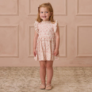 A young girl smiles in her Noralee Baby Girls' and Girls' Alice Dress, a pink floral design with ruffled sleeves, against a beige paneled wall. The dress boasts intricate embroidered lace details that enhance the whimsy of her charming ensemble.