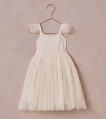 The Girls' Poppy Dress by Noralee is an elegant ivory dress with a tulle skirt and feathered shoulder straps, beautifully adorned with floral embroidery. It hangs gracefully on a rose gold hanger against a textured beige background.