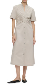 A person is wearing the Noor Dress, a Callas Milano beige midi shirt dress with short sleeves, front buttons, and a flattering fit and flare design with gathered detail at the chest. They are also wearing black flat shoes.