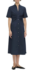 A woman wears the Callas Milano Noor Dress, a navy midi-length shirt dress with short sleeves and front-gathered detail, perfectly paired with black slip-on shoes. The flattering fit and flare design effortlessly complements her style.
