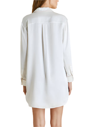 A woman is seen from the back wearing the Olivia von Halle Poppy Silk Nightshirt, a long-sleeved, white piece that falls to mid-thigh—ideal for luxury sleepwear or as an elegant bridal trousseau addition.