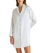A woman wears the Poppy Silk Nightshirt by Olivia von Halle—a long-sleeve, white satin shirt with buttons and a chest pocket. This relaxed-fit nightshirt is perfect for luxury sleepwear or a bridal trousseau.