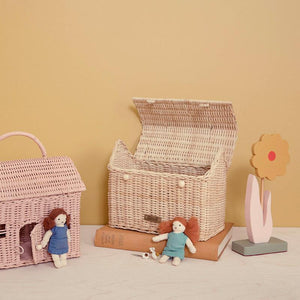 An open wicker basket on a light surface against a beige background contains two small rag dolls, an Olli Ella Casa Clutch in straw, books, and a wooden flower decoration.