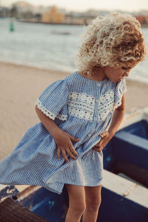 A curly-haired child wears the Louise Misha Girls' Morlina Dress, a blue striped, textured cotton dress with lace details, while standing near a boat on the beach.