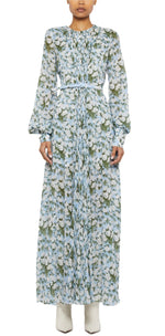 A person wearing the Prabal Gurung Huma Maxi Dress—a long-sleeved, light blue floral, floor-length dress with a flowy silhouette and belted waist—paired with white boots stands against a plain white background.