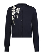 The Lela Rose Raffia Embroidered Knit Long Sleeve Cardigan features a navy blue knit fabric, button-up front, and white floral embroidery on the left shoulder and chest.