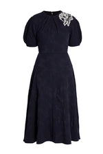 The Erdem Short Sleeve Midi Dress features a knee-length navy jacquard design with short puff sleeves, subtle textured pattern, and a white floral applique accenting one shoulder.