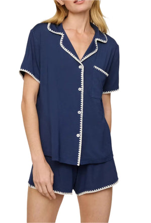 A woman wears the Eberjey Frida Relaxed Short PJ Set: a navy short-sleeve, white-trim button-up top with artisanal whipstitch and matching boyfriend-fit shorts.