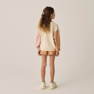 A young girl with curly hair, wearing Rylee & Cru Girls' Sweat Shorts and a matching beige and peach french terry sweatshirt with white sneakers, stands facing away against a plain background.