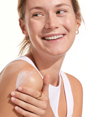 A woman in a white tank top smiles while applying Supergoop Play Everyday Lotion SPF 50 with Sunflower Extract (2.4 oz) to her shoulder.