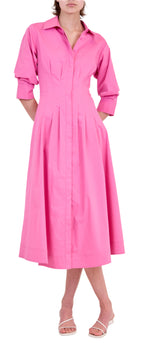 A woman wears the Signature Jazz Dress by Simkhai, a long-sleeved pink cotton poplin midi with pintucks, pockets, and an A-line silhouette, paired with white heeled sandals.