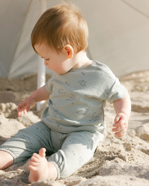 A baby wearing the Quincy Mae Baby Boys' Jersey Tee & Pant Set with dino print sits on sandy ground beneath a white umbrella, gazing downward and to the side.