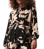 A person wearing the Kasey Tunic by Figue, a black and beige floral-patterned blouse with long sleeves, waist tie detail, and a stylish boat neckline.