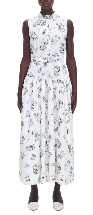 A person in an Arden Dress by Adam Lippes, a sleeveless, long white floral dress made of Italian cotton poplin with a subtle drop waist, stands against a plain background wearing white shoes.