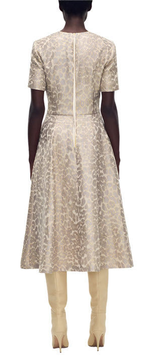 Someone is seen from behind wearing the Eloise Dress Leopard Jacquard by Adam Lippes, a knee-length, short-sleeve dress in a bold leopard pattern with a back zipper, paired with knee-high beige boots.