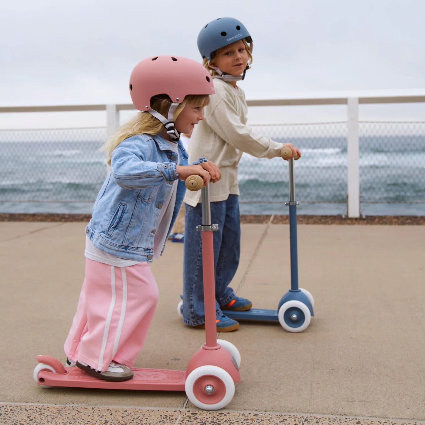 Two children ride Banwood Eco Scooters along a seaside path, both wearing helmets and casual clothes—the girl on a pink recycled-plastic scooter, the boy on a blue one.