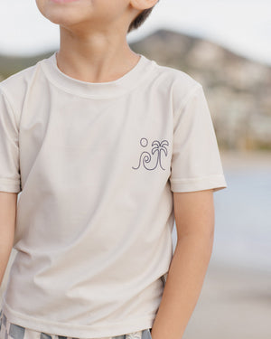 A child wears the Rylee & Cru Boys' Rashguard in light beige with a palm tree and sun graphic, standing outdoors near hills and water—ideal for beach or pool play with UPF 50+ protection.