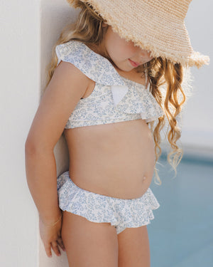 A young child in a Rylee & Cru Girls' Skirted Bikini with an asymmetrical ruffle top and straw hat stands by a white wall near a pool, face partly shaded. UPF 50+ fabric offers sun-safe style for all-day play.