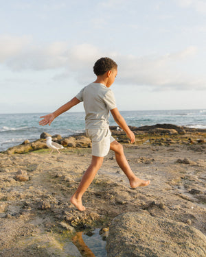 A child wears the Rylee & Cru Baby and Kids' Short-Sleeve Rashguard, walking barefoot on rocky ground by the ocean under a cloudy sky, with a seagull in the background.