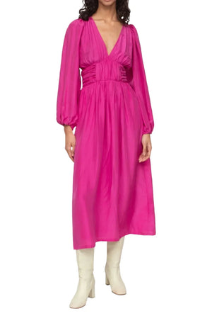 A person wearing the Sea NY Fabiola Dress in bright pink silk habotai, featuring a V-neckline, gathered waist, and long sleeves, paired with white knee-high boots.
