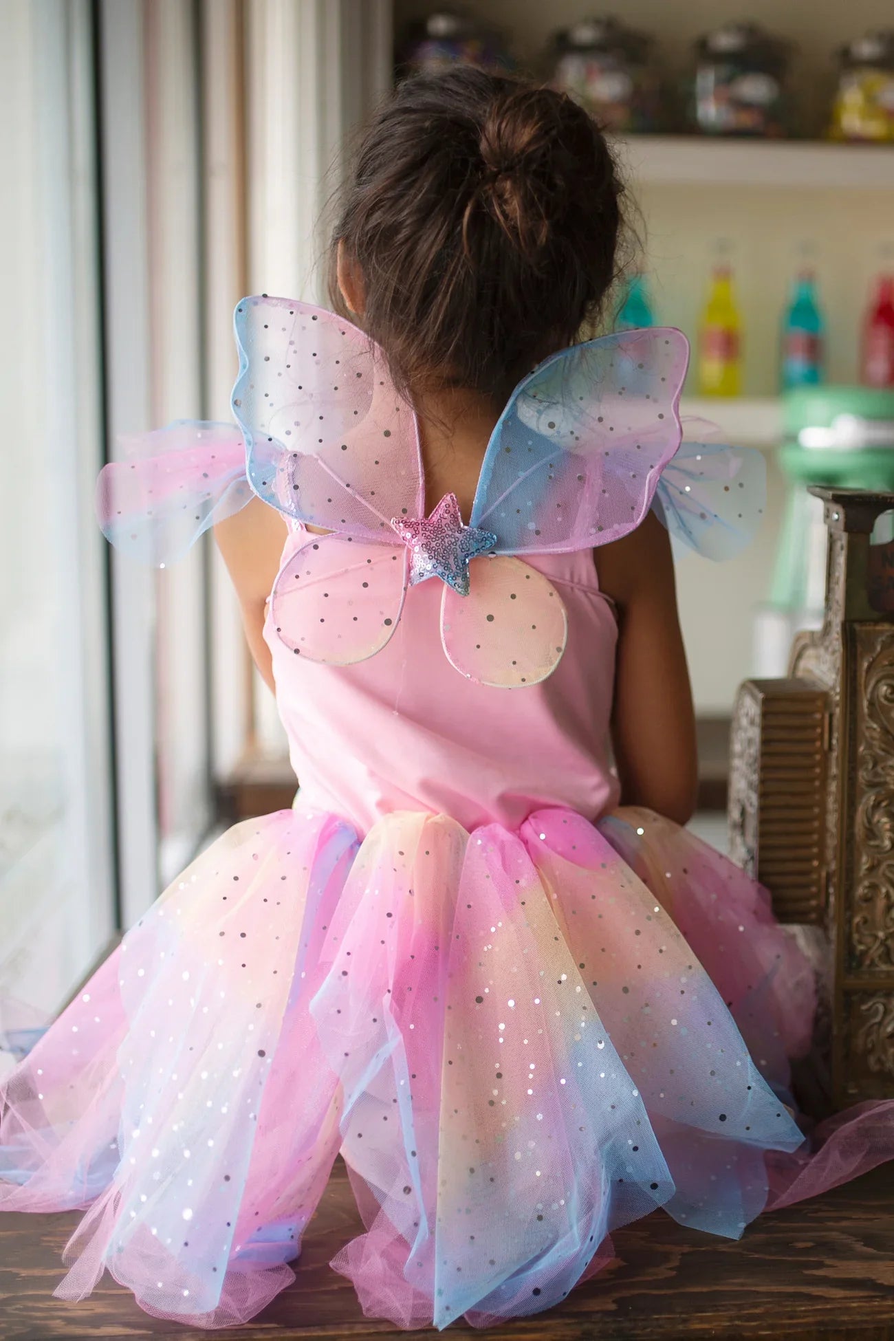 Indoors, a child sits facing away in the Rainbow Fairy Dress & Wings by Great Pretenders, featuring a pink dress with shimmery organza, multicolored tulle, and butterfly wings.