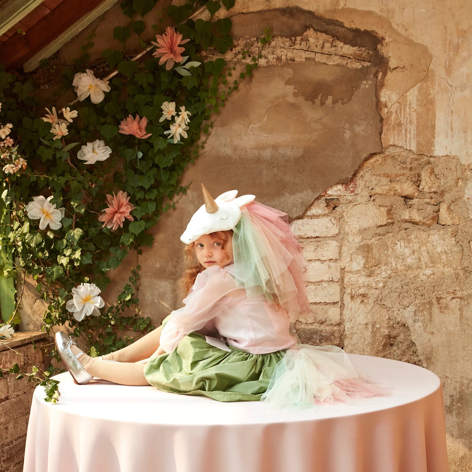 A child in a Meri Meri Rainbow Unicorn Costume sits on a round table with a pink tablecloth, surrounded by greenery and flowers against a rustic wall.