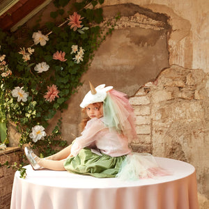 A child in a Meri Meri Rainbow Unicorn Costume sits on a round table with a pink tablecloth, surrounded by greenery and flowers against a rustic wall.