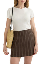 A woman wearing a white short-sleeve top, the POSSE Alice Pinstripe Skirt, and carrying a woven shoulder bag.