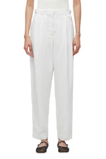 Against a plain backdrop, an individual showcases the 3.1 Phillip Lim Double Pleated Tapered Trouser in soft poplin. The relaxed white fabric elegantly drapes over black shoes, merging comfort with sophistication.