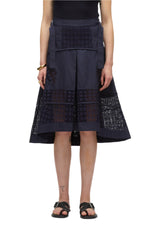 Dressed stylishly, a person sports a black sleeveless top with the Broderie Anglaise Grid Skirt by 3.1 Phillip Lim. Paired with black sandals and accessorized with a bracelet, they prominently display an ankle tattoo against the navy midi skirt featuring lace details that flow into an A-line silhouette.