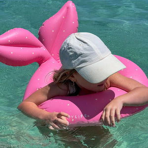 A confident child floats on a Sunnylife Kiddy Pool Ring, resembling a pink mermaid tail in clear blue waters, their white cap gleaming under the sun.
