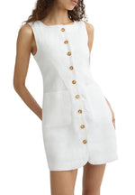 A woman wears the POSSE Emma Mini Dress—a sleeveless white linen mini dress featuring a high neckline, large front pockets, brown center buttons, and an adjustable back buckle for the perfect fit.