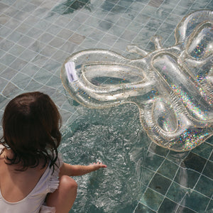 A child in a white swimsuit touches the water in a tiled pool beside a Sunnylife Kids' Lie-On Float Cotton Candy Glitter.